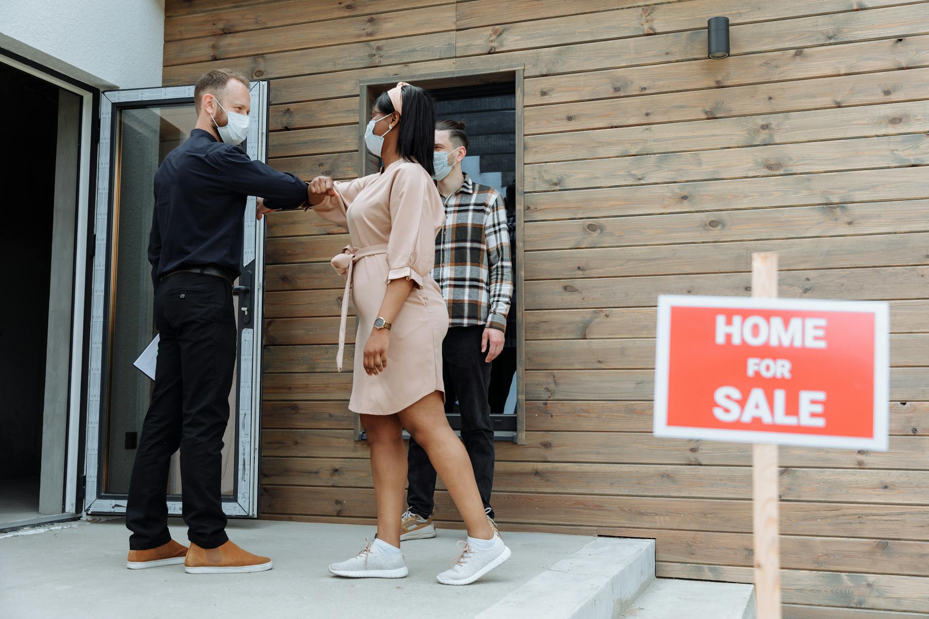 Masked real estate agent greets clients with elbow bump outside a home for sale.
